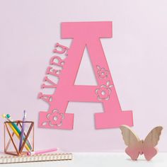 a pink wooden letter sitting on top of a desk next to a cup and pencils