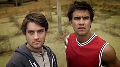 two young men standing next to each other in front of a dirt field and fence