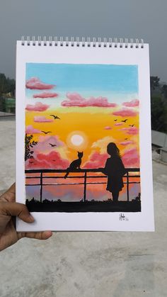 a person holding up a painting in front of a sunset with a cat sitting on a fence