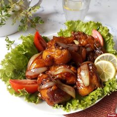 chicken with onions and tomatoes on a plate next to a glass of water, lemon wedges and some herbs