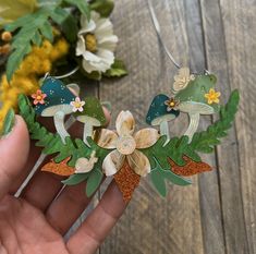 a hand is holding an ornament made out of paper flowers and leaves on a wooden table