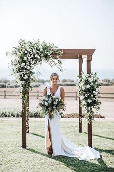 a woman in a white dress standing under an arch with greenery and flowers on it