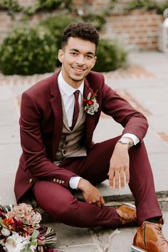 a man in a suit and tie sitting on the ground next to some flowers, smiling