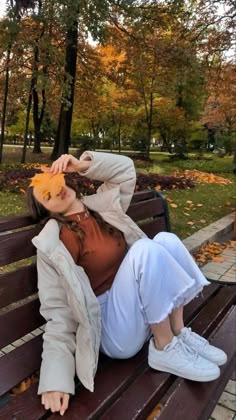 a woman sitting on top of a wooden bench in front of some trees and leaves