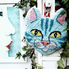 a crocheted blue cat head hanging on a wall next to some green plants