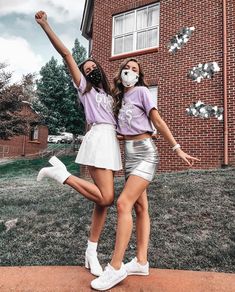 two girls wearing face masks standing in front of a brick building with their arms up