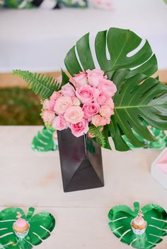 a vase with pink flowers and greenery on a table in front of palm leaves