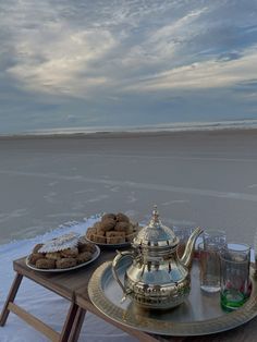 two trays with pastries and drinks sit on a table at the beach in front of an ocean