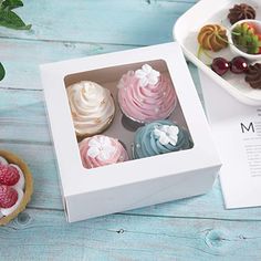 four cupcakes in a white box on a blue wooden table next to a bowl of fruit