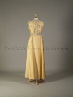 Pierre Balmain Haute Couture evening dress from French 1960-1970. Made from yellow silk satin peau de soie ground, yellow dome caps, embroidered  embroidery with bead, pearl, rhinestones, cream cord loops, white plastic lilies flower, light yellow braid trim,  and dome lucite button. Sleeveless floor length A line gown in scoop necked style.#Balmain #HauteCouture #Vintage #Fashion House of Balmain. Dress Creator, Kent State University, Kent State, Evening Formal