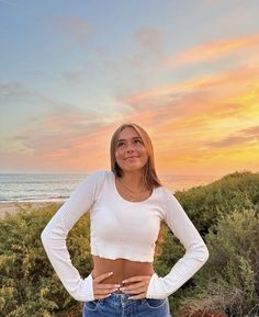 a woman standing in front of the ocean with her hands on her hips looking at the camera