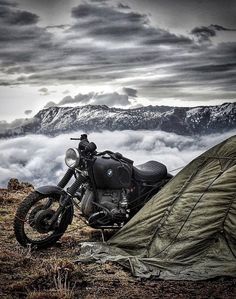 a motorcycle parked next to a tent on top of a hill with mountains in the background