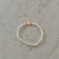 a white beaded bracelet with pink beads and a gold ball on it, sitting on a marble surface