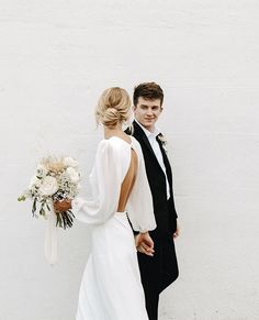 a man and woman standing next to each other in front of a white wall holding hands