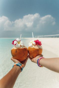 two people holding up coconuts in front of the ocean with white sand and clear blue water