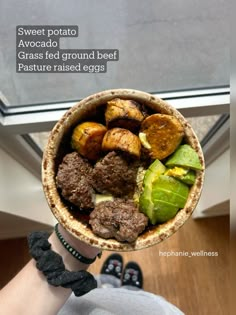 a person holding up a bowl filled with meat and veggies in front of a window