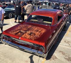 an old red car parked in a parking lot with other cars and people standing around