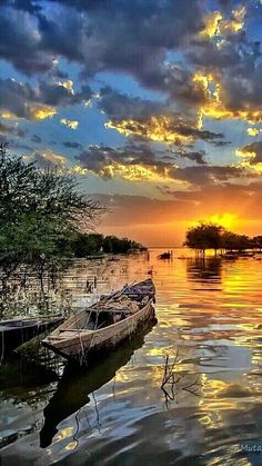 a boat is sitting in the middle of water at sunset with clouds and trees around it