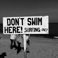 black and white photograph of people standing next to a sign that says don't swim here surfing only