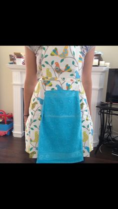 a woman is wearing an apron with birds on the front and blue bottom, while standing next to a fireplace