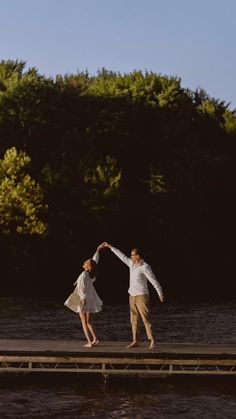 two people are dancing on a dock by the water with trees in the back ground