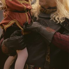 a man with long blonde hair holding a small child in his arms while wearing medieval clothing