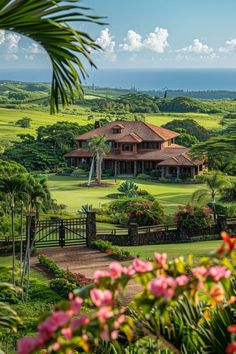 a large house surrounded by lush green trees and flowers in the foreground, with an ocean view behind it