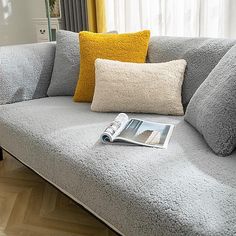 a grey couch with yellow pillows and a magazine on the floor next to it in front of a window