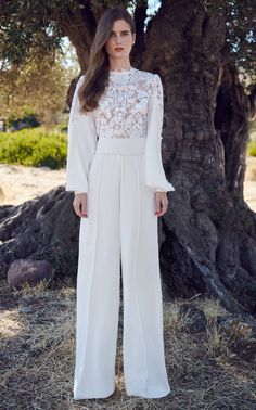 a woman standing in front of a tree wearing white pants and a top with lace on it