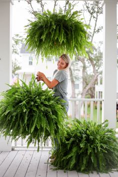 a woman standing on a porch next to two large green plants hanging from the ceiling