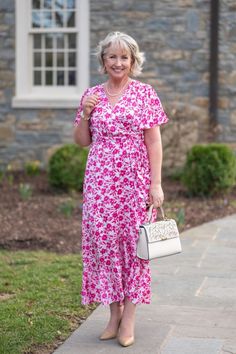 a woman in a pink floral dress holding a white purse and smiling at the camera