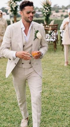 a man in a suit and sunglasses is walking through the grass with an orange drink