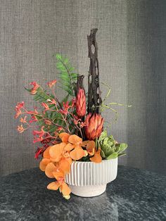 an arrangement of flowers in a white bowl on a table with grey wall behind it