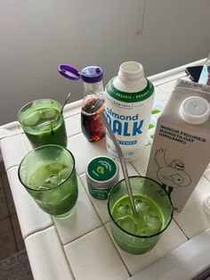 three glasses filled with green liquid sitting on top of a counter next to bottles and spoons