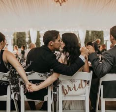 two people sitting on chairs kissing each other in front of a tent with white walls