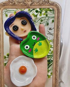 a hand holding a small doll with an egg in it's bowl next to a mirror