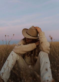 a woman sitting in the middle of a field wearing a hat and holding her hands to her face