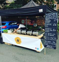 a man preparing food under a black tent on the street next to a blue car