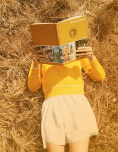 a woman in yellow shirt and white shorts reading a book while standing on dry grass