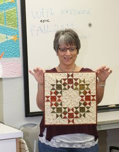 a woman holding up a piece of quilt in front of a white board with writing on it