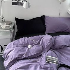 an unmade bed with purple sheets, black pillows and a white lamp on the side