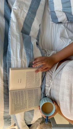 a woman laying in bed reading a book and holding a cup of coffee with both hands