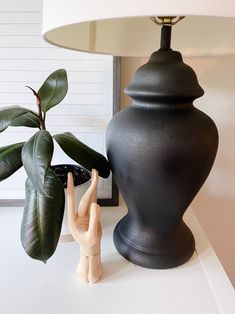 a black vase sitting next to a lamp on top of a white table with a plant in it