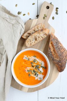 a bowl of carrot soup with bread and seeds on a cutting board next to it