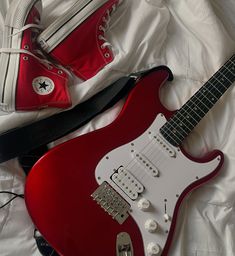 a red electric guitar sitting on top of a bed next to a pair of converse shoes