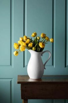 a white vase filled with yellow flowers sitting on top of a wooden table next to a blue wall