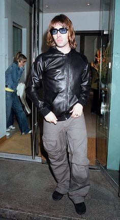 a man in sunglasses and a leather jacket is standing on the sidewalk with his hands in his pockets