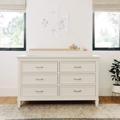 a white dresser with two windows and a potted plant in front of the dresser