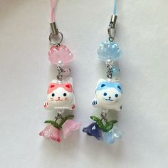 two charms with cats and flowers are hanging on a white surface, one has a pink ribbon