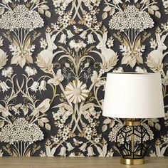 a lamp sitting on top of a wooden table next to a wall covered in flowers